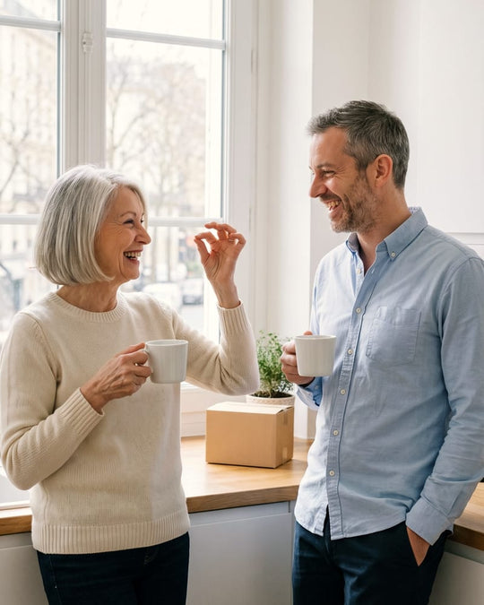 Une mère et son fils adulte rient, une tasse à la main, dans une cuisine lumineuse.